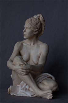 A sculpture in clay of a woman sitting looking to the left holding her left leg close to her body. There is a cloth covering her right leg.